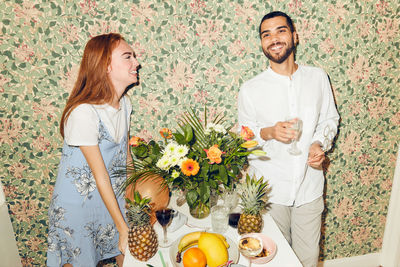 Smiling young friends standing by table against wallpaper during dinner party at home