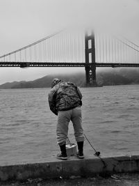 Rear view of man standing on retaining wall near golden gate bridge at harbor