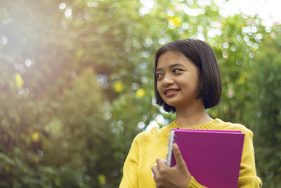 Smiling girl holding book against trees