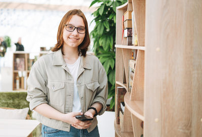 Young brunette teenager girl student in glasses with mobile phone at modern library, public place