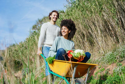 Woman sitting in wheelbarrow, holding fresh vegetables, man pushing her