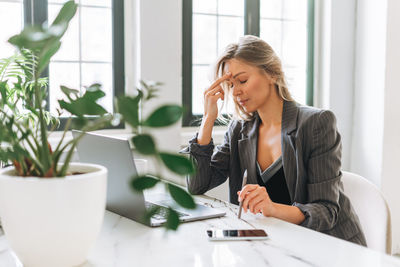 Young tired blonde woman with long hair in stylish grey suit working at laptop in bright office