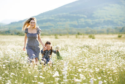 Cute young woman holding hands and play with child girl in chamomile field with blooming flower