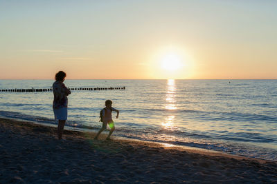 Rear view of children standing on beach against sky during sunset