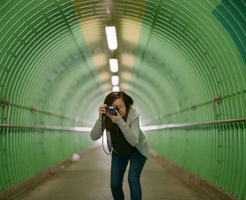 Portrait of young woman photographing with camera while standing in green tunnel