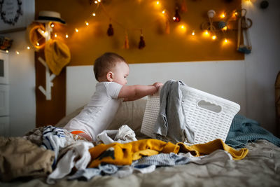 Funny european chubby kid pulling out clothes from laundry basket on bed in cozy bedroom,