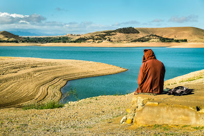 Rear view of man sitting on land by lake against sky