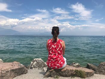 Rear view of woman sitting on rock at beach against sky