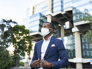 Serious black male entrepreneur in formal suit and medical mask standing in downtown waiting for meeting looking away