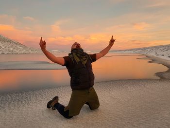 Full length of man screaming while kneeling at beach against sky during sunset