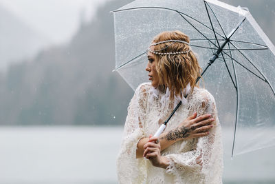 Beautiful young woman bride in a boho dress and with an umbrella stands in the rain in nature