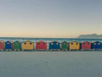 A line of small colorful beach houses on muizenberg beach in front of the blue ocean during sunset.