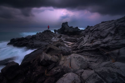 Distant view of man standing on rock formation against cloudy sky at dusk