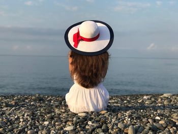 Rear view of woman wearing hat sitting on pebbles at beach against sky during sunset