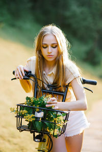 Young beautiful woman standing near yellow bicycle with wicker basket full of flowers in forest