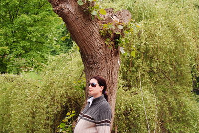 Pregnant woman wearing sunglasses and jacket standing against plants and trees