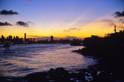 Silhouette buildings by sea against sky at sunset