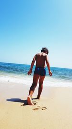 Rear view of shirtless boy walking at beach against blue sky