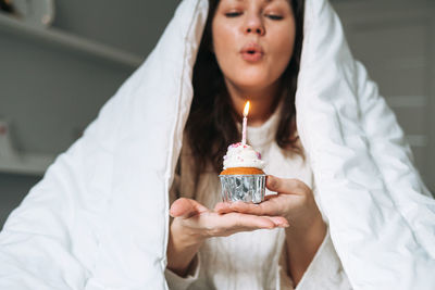 Young woman body positive in white blanket with festive cupcake with candle in hands on bed at home 
