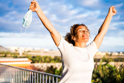 Smiling woman holding mask while standing with arms raised