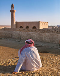 Rear view of man in traditional clothing sitting by built structure