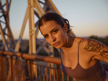 Trendy young female with tattooed arms leaning on metal fence and looking at camera