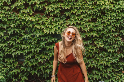 Cheerful young woman wearing sunglasses while standing against plants