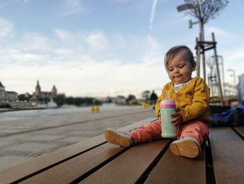 Cute girl sitting on bench against sky