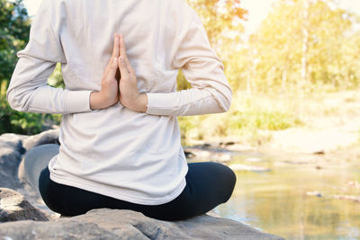 Rear view midsection of woman with hands clasped while practicing lotus position on rock by river