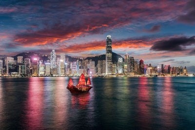 Illuminated buildings by sea in city against sky at sunset