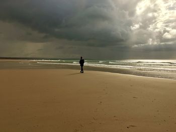 Full length of a person walking alone on beach against dramatic sky
