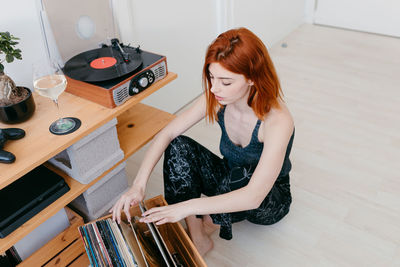 Young female selecting vinyl record from wooden container while sitting against vintage turntable in house room