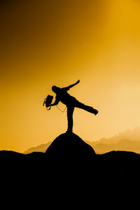 Silhouette man holding camera while standing on rock against sky during sunset