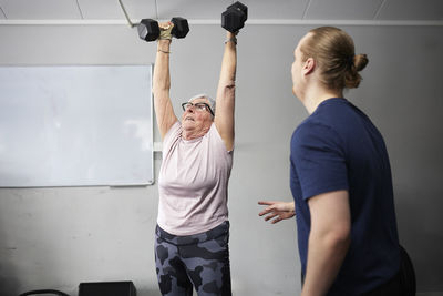 Fitness instructor standing near senior woman exercising with dumbbells in gym