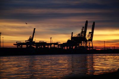 Silhouette cranes at commercial dock against sky during sunset