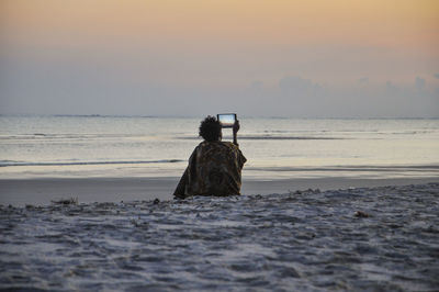 Rear view of woman using digital tablet at beach against sky during sunset