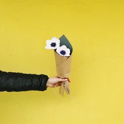 Cropped hand of woman holding wrapped flowers against yellow wall