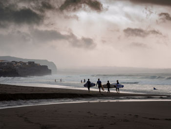 Full length of friends with surfboards walking at beach against sky during sunset