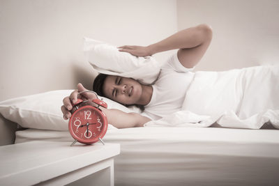 Young man pressing head with pillow while closing alarm clock