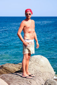 Shirtless mid adult man wearing sunglasses standing at rocky beach against clear sky