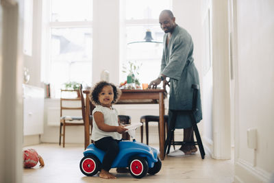 Portrait of girl sitting on toy car with father standing in background at dining room