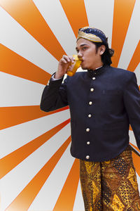 Young man wearing traditional clothing while drinking fresh jamu against pattern wall