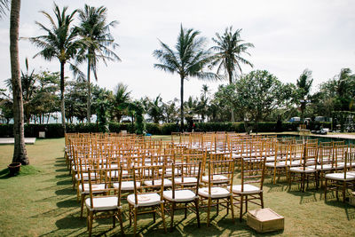 Empty chairs and table by palm trees against sky