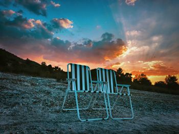 Empty chairs at beach against sky during sunset