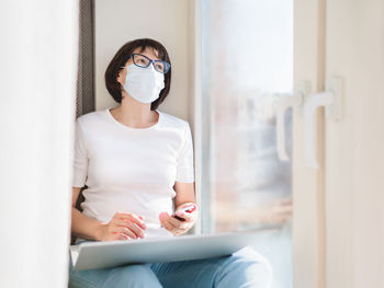 Woman in medical protective mask works remotely from home. she sits with smartphone and laptop. 