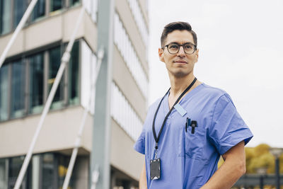 Confident male doctor wearing blue scrubs looking away in front of hospital building