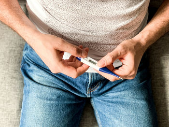 Man is checking temperature with digital thermometer, sitting on gray sofa with thermometer in hands