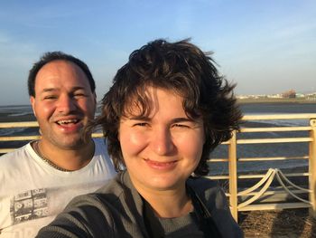 Portrait of smiling couple standing at beach against sky during sunset