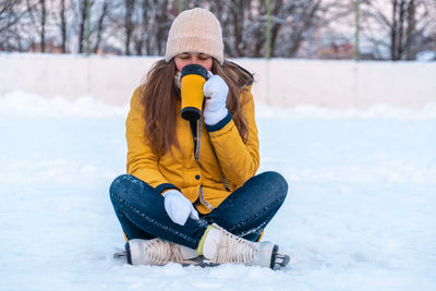 Portrait of young woman in ice skates sitting on the snow and drinking coffee