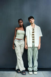 Full body of multiracial man and woman in loose pants and simple tops standing close and holding hands on gray background